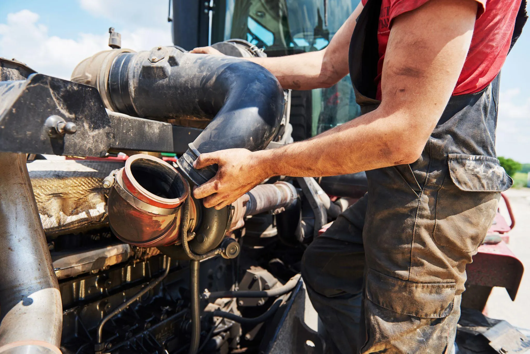 A worker in dirty overalls refitting a large rubber intake hose on a diesel engine