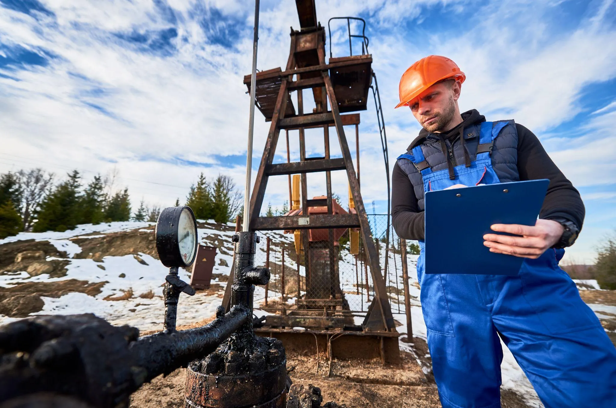 A worker in a hard hat writing on a clipboard beside a wellhead pressure gauge, with a pump jack behind him