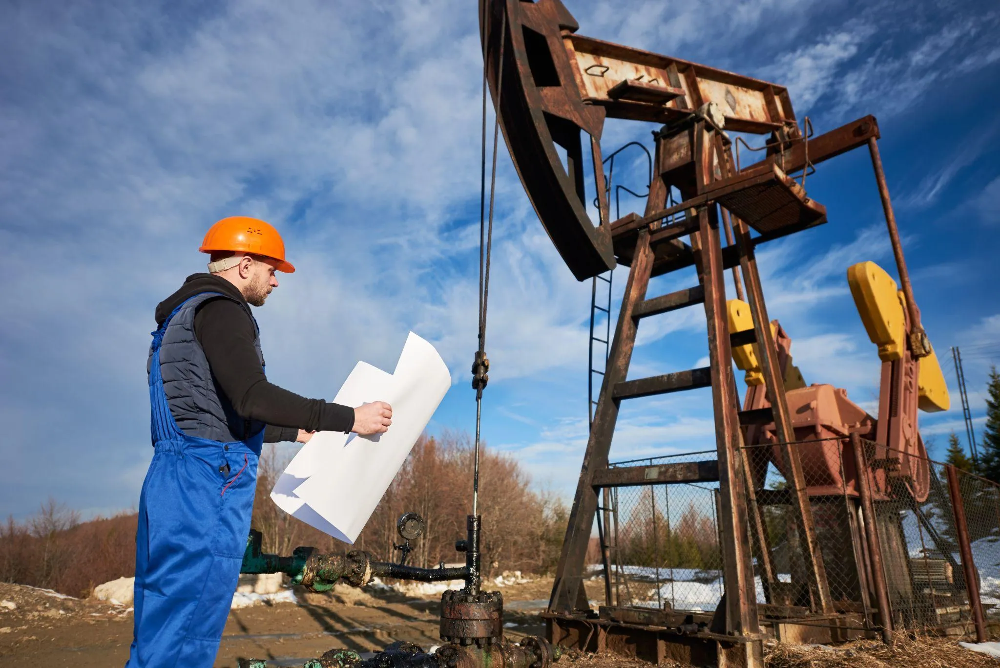 A worker in a hard hat reading a large sheet of plans beside a wellhead, with a pump jack behind him